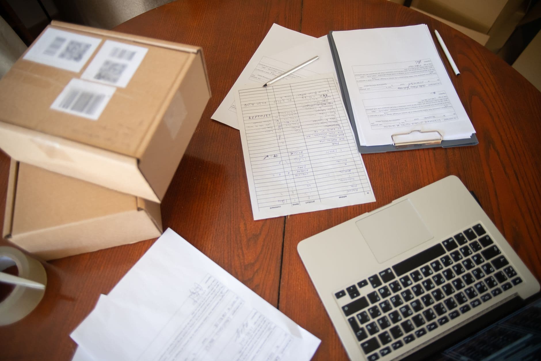 Shipping paperwork, a packing list and a laptop on a desk beside sealed cartons
